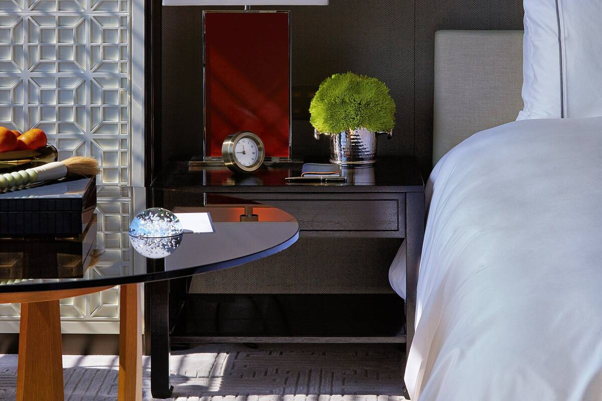 A hotel room with a bed, a nightstand with a lamp, a clock, and a small table with a vase of greenery.