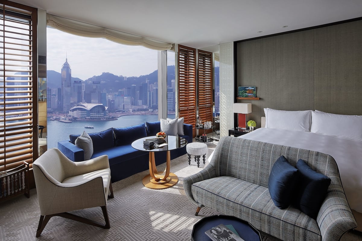 A hotel room with a large bed, a sofa, a chair, a small table, and a view of a cityscape.