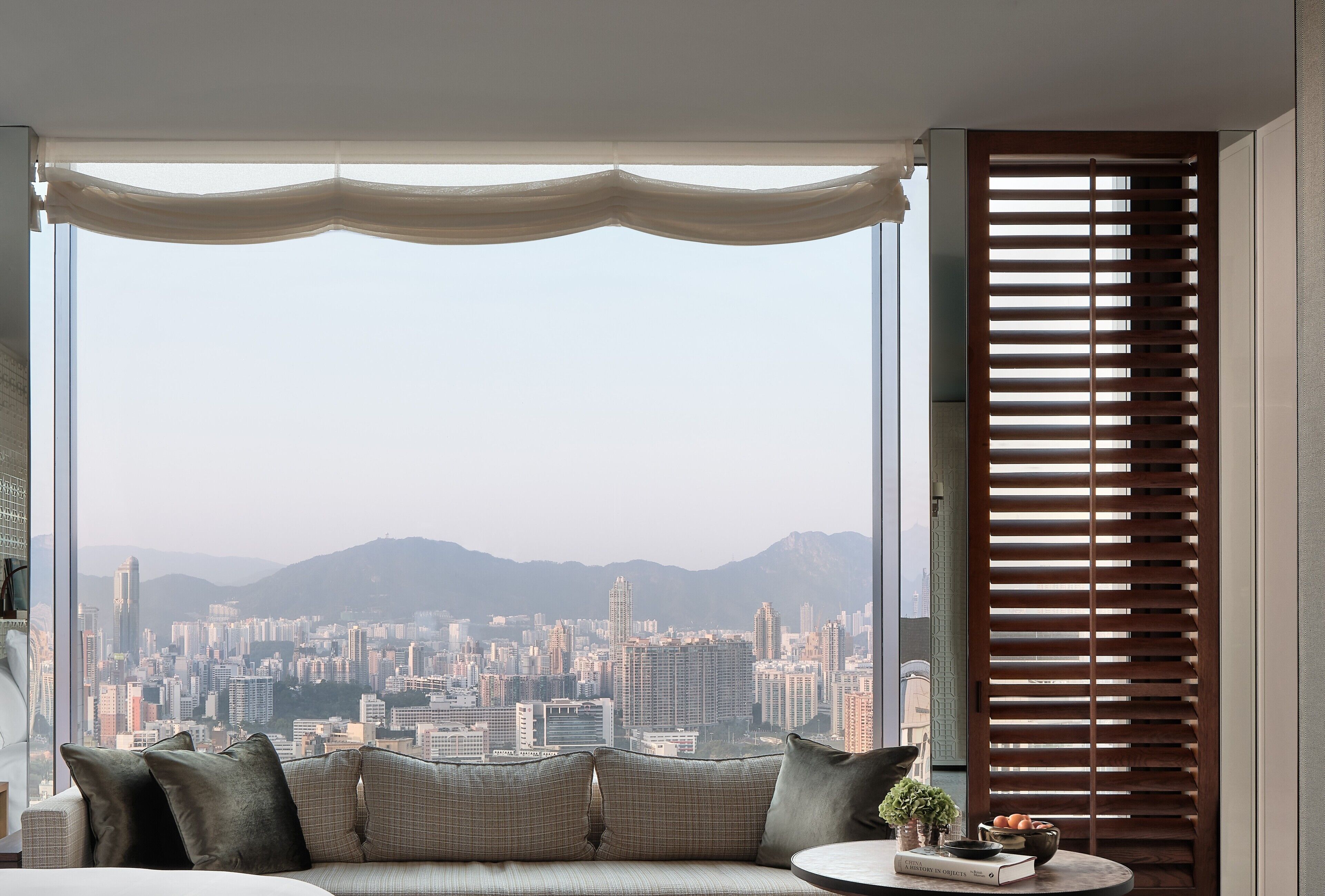 A modern living room with a large window offering a city view, a sofa with cushions, and a small table with decorative items.