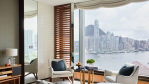 A hotel room with a large window offering a city view, a desk with a lamp, two chairs, and a bed.