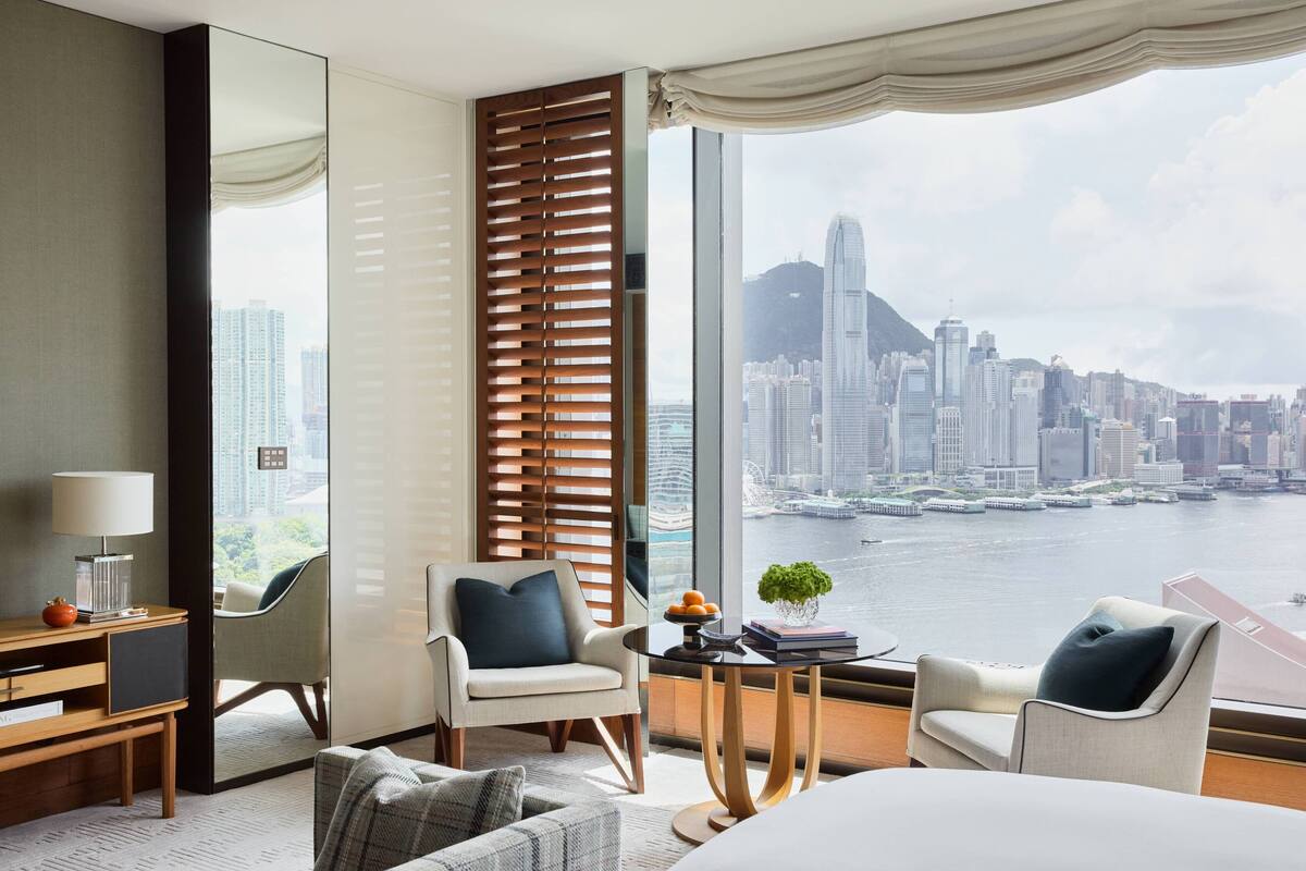 A hotel room with a large window offering a city view, a desk with a lamp, two chairs, and a bed.