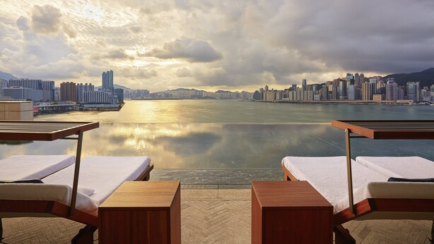 A waterfront hotel balcony with two sun loungers, a view of a city skyline across a body of water, and a cloudy sky.
