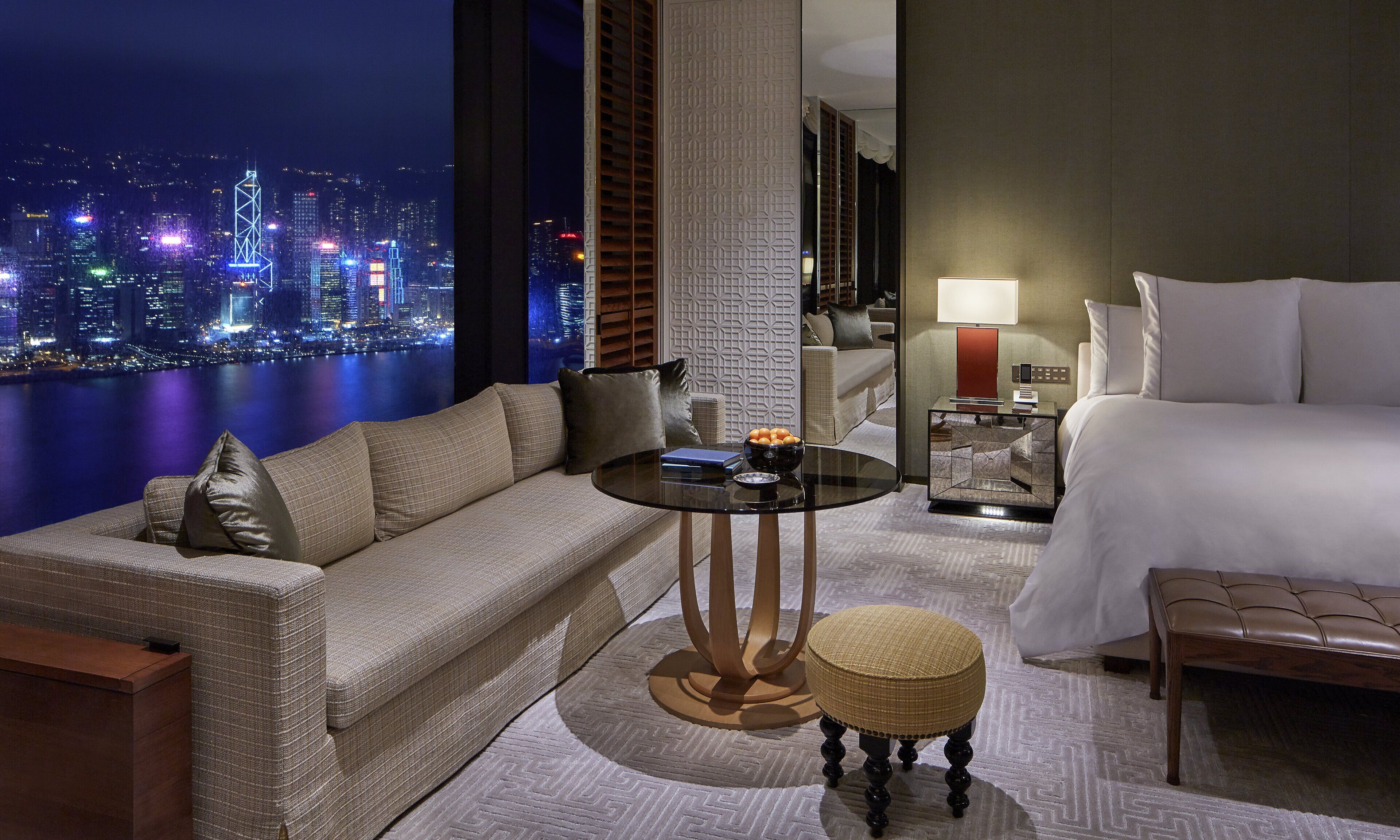 A hotel room with a sofa, a small round table, a bedside table with a lamp, a bed, and a view of a cityscape at night.