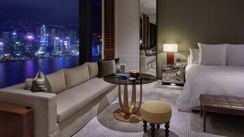 A hotel room with a sofa, a small round table, a bedside table with a lamp, a bed, and a view of a cityscape at night.