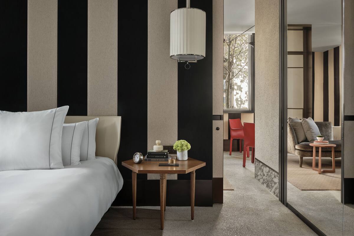 A modern hotel room with a bed, a small wooden table with books and a clock, a red chair, and a large window with a view of the outdoors.