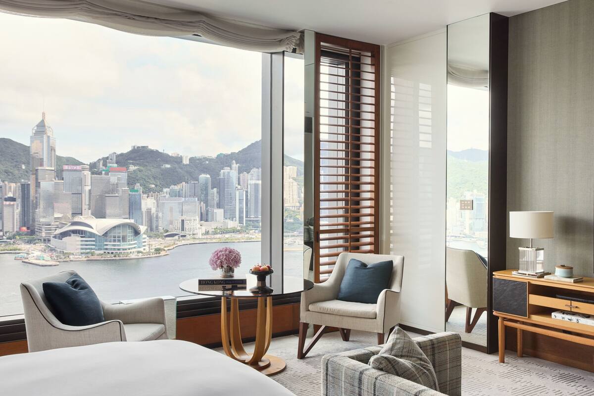 A modern hotel room with a large window offering a city view, a comfortable seating area, and a small side table with a lamp and books.