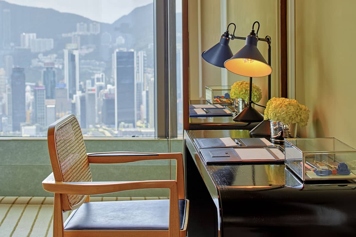 A hotel room with a view of a cityscape, a desk with a lamp, a chair, and a vase with flowers.