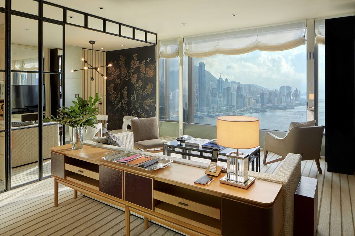 A modern hotel room with a large desk, a comfortable seating area, a TV, and a city view.