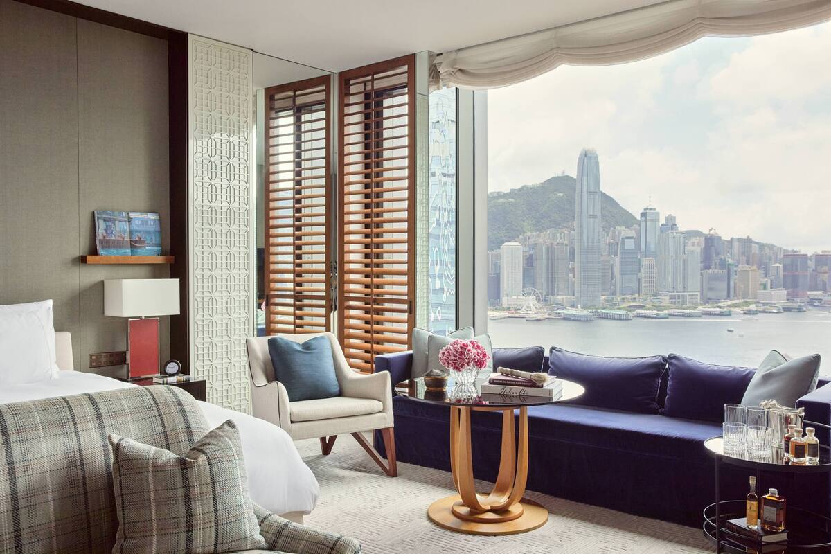 A hotel room with a bed, a sofa, a table with a vase of flowers, and a view of a cityscape.