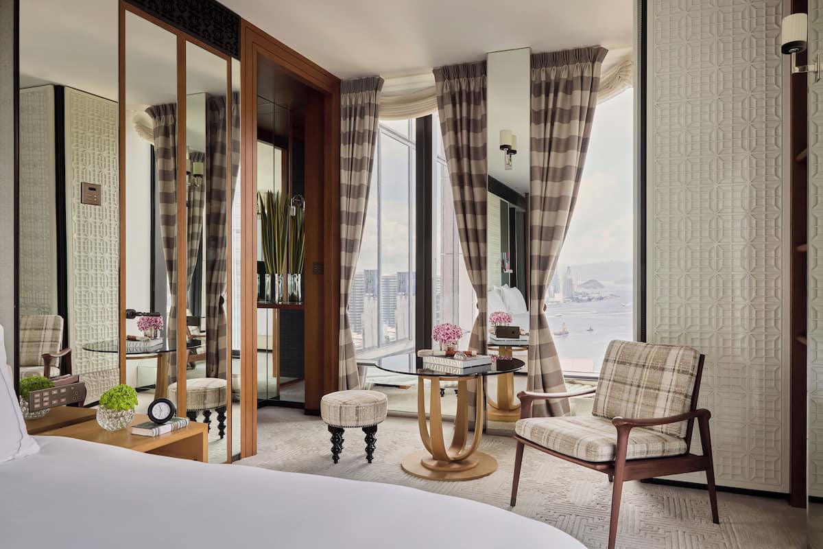 A hotel room with a large bed, a round table, a chair, a small stool, and a view of the city.