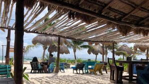 Ein Sitzbereich am Strand mit Reetdachschirmen, Holztischen und -stühlen.
