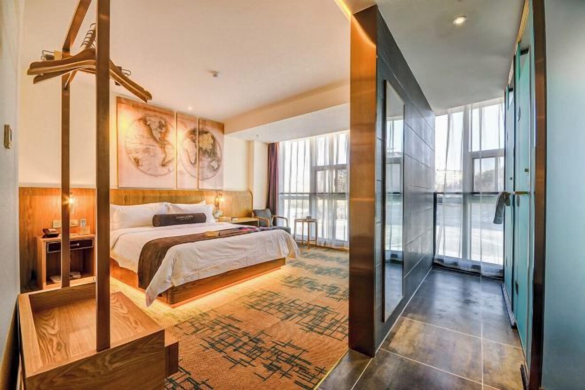 A modern hotel room with a large bed, a wooden side table, a large window, and a view of the outdoors.