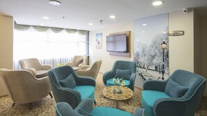 Et hotel lobby med et siddeområde med lænestole og sofaer, et sofabord med blomster, et fjernsyn ophængt på væggen og et billede der viser et sneklædt gadesceneri.