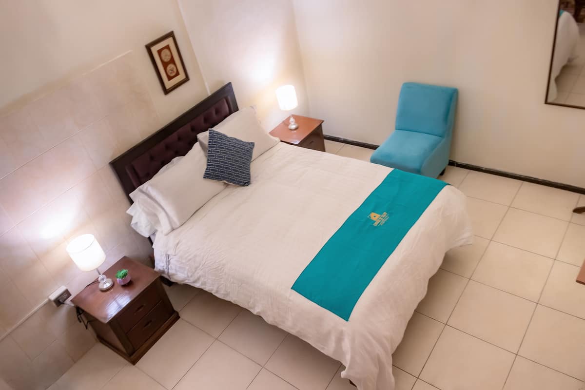 A neatly made bed with a white and teal bedspread, a wooden nightstand with a lamp, and a blue chair in a hotel room.