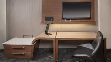 A hotel room with a wooden desk, a black office chair, a flat-screen TV, and a small speaker.