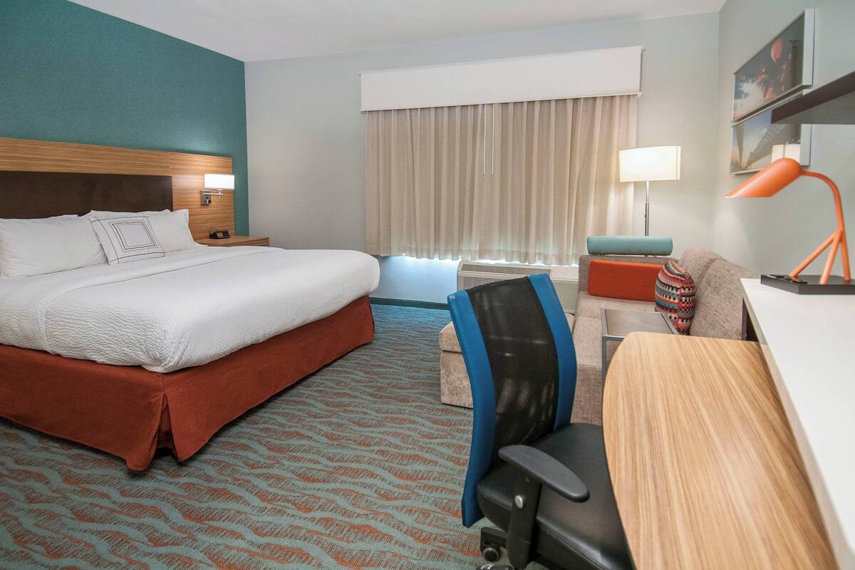 Een hotelkamer met een groot bed, een bureau met stoel, een bureaulamp, een raam met gordijnen, en een wanddecoratie.