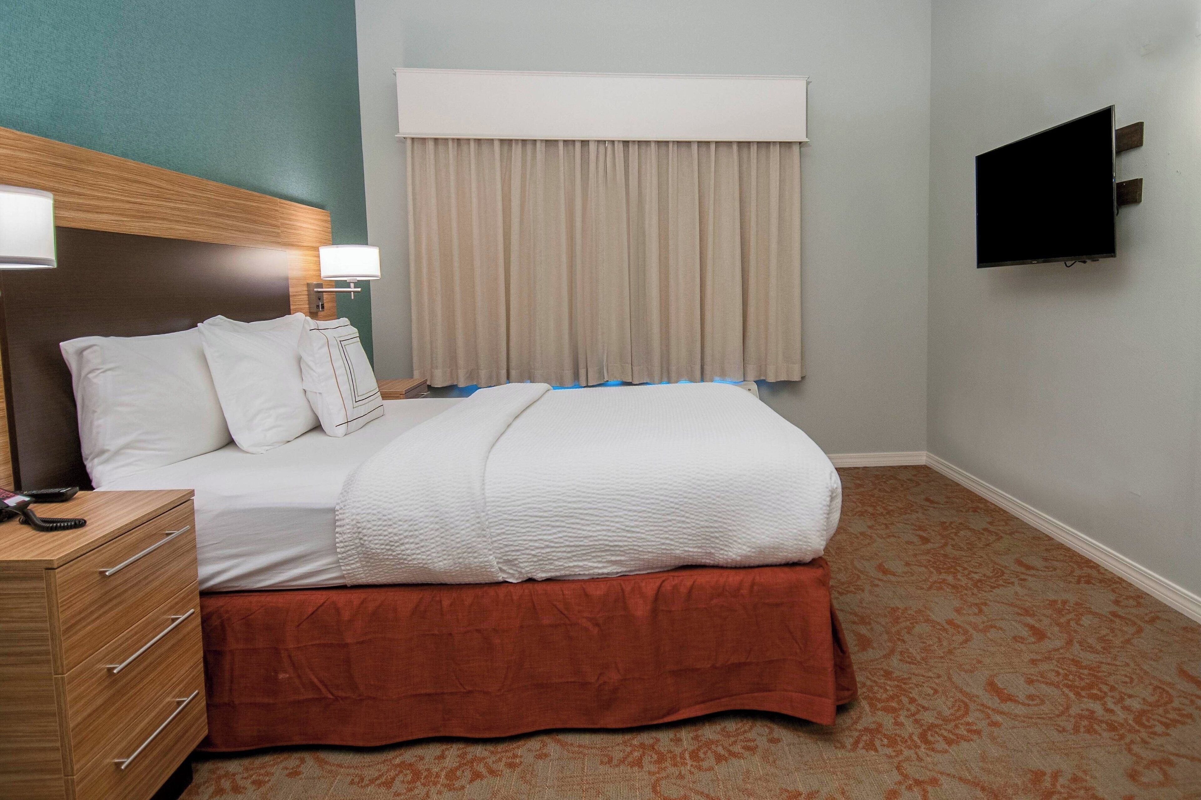 Une chambre d’hôtel avec un lit, une commode en bois, une télévision et une fenêtre avec des rideaux.