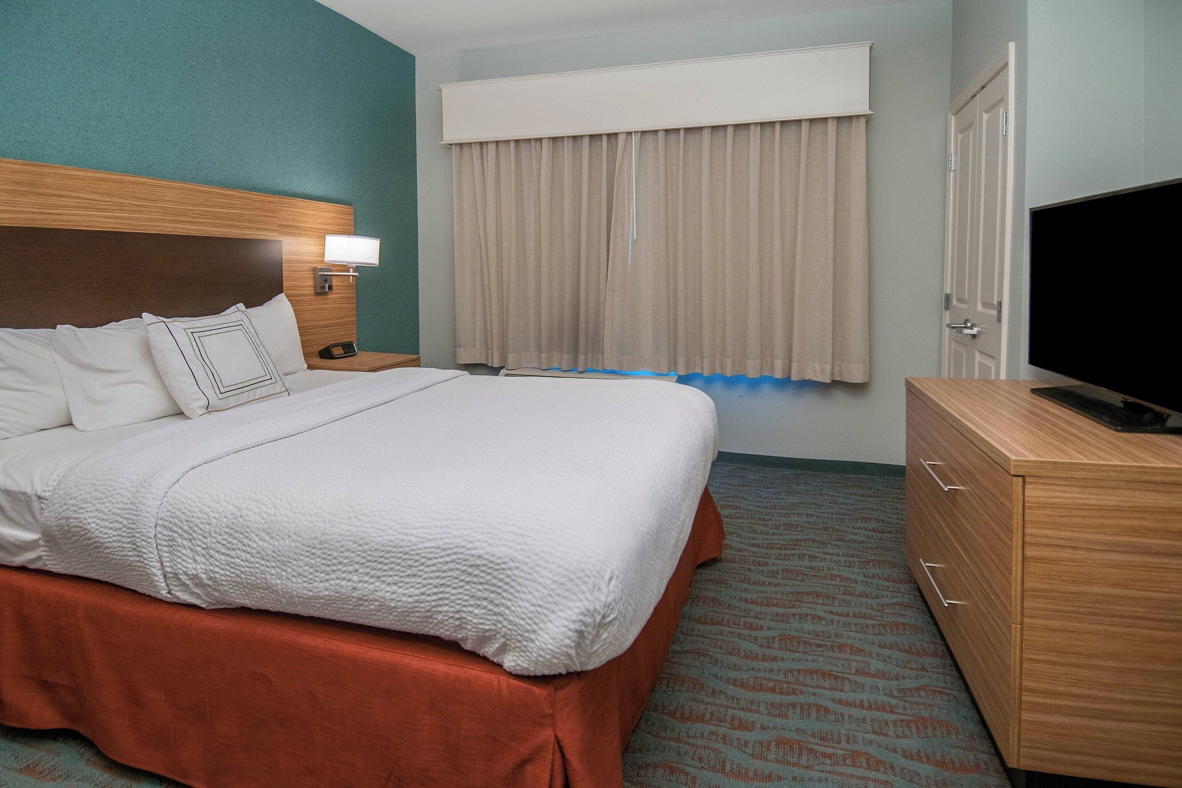 Quarto de hotel com uma cama grande, cabeceira de madeira, mesa de cabeceira com abajur, televisão e guarda-roupa.