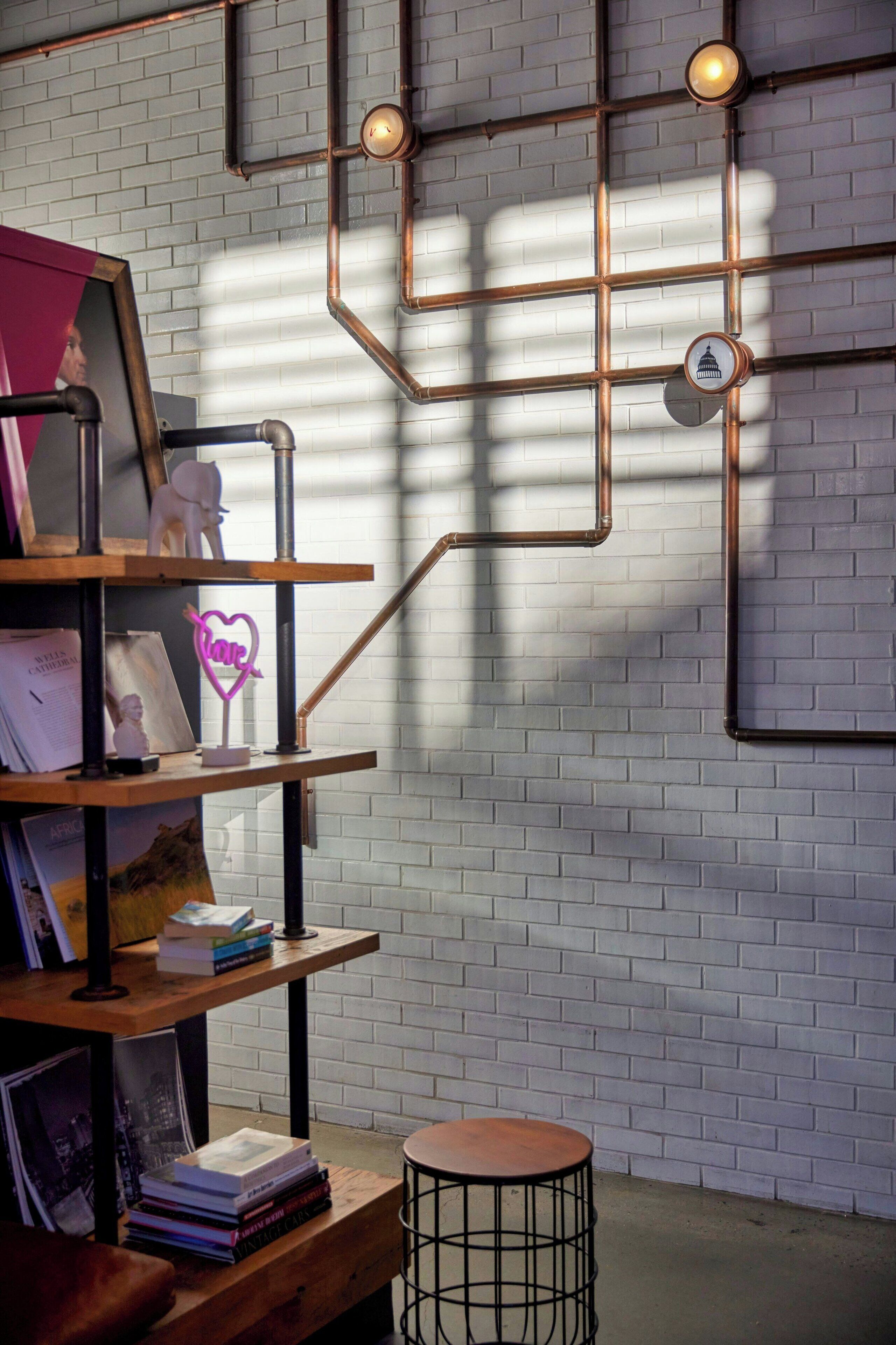 Een kamer met een bakstenen muur, een houten plank met boeken en een neonbord, een rond krukje en een wandlamp.