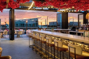 A rooftop bar with a long counter, bar stools, and a view of the city skyline at sunset.