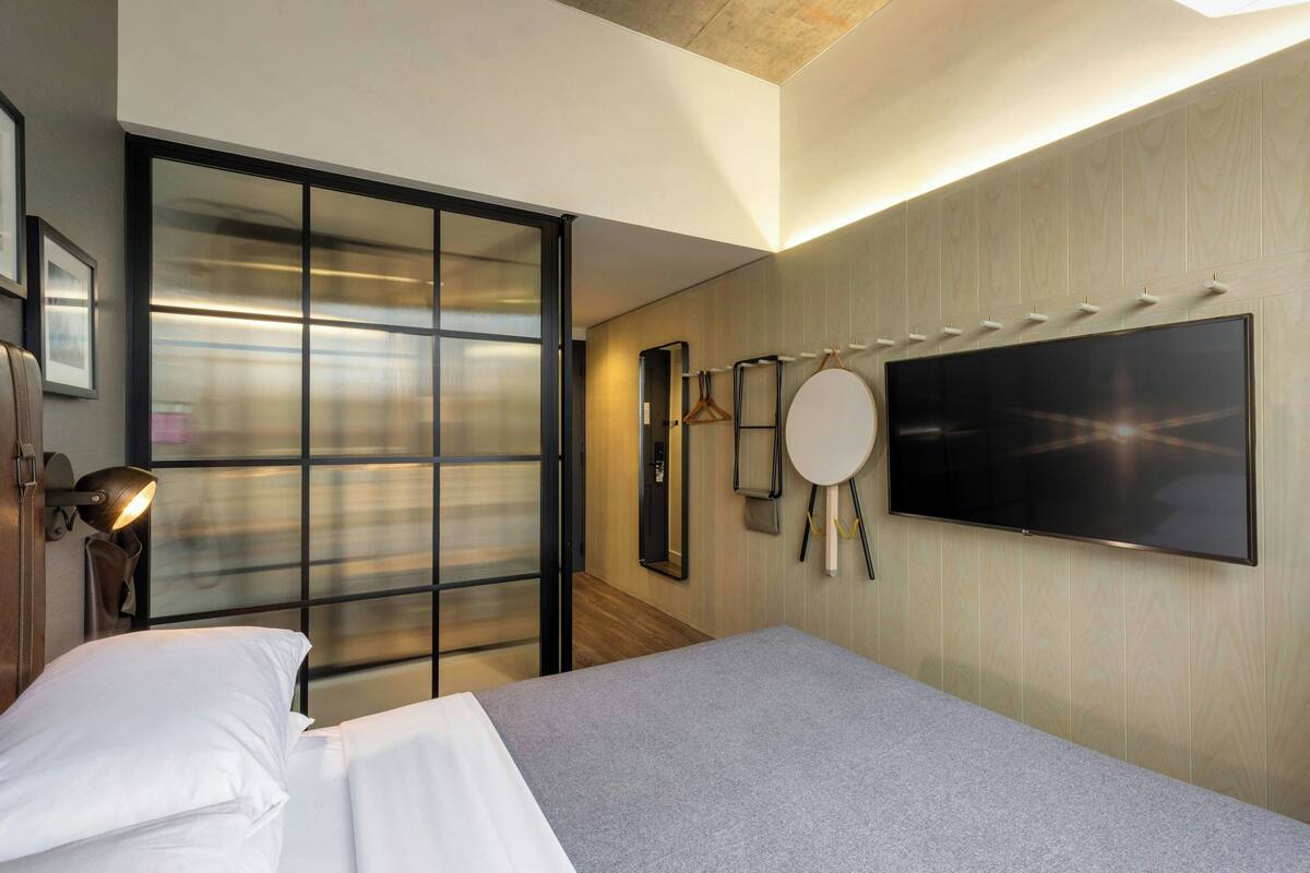A modern hotel room with a bed, a TV mounted on the wall, a sliding door, and a round mirror.