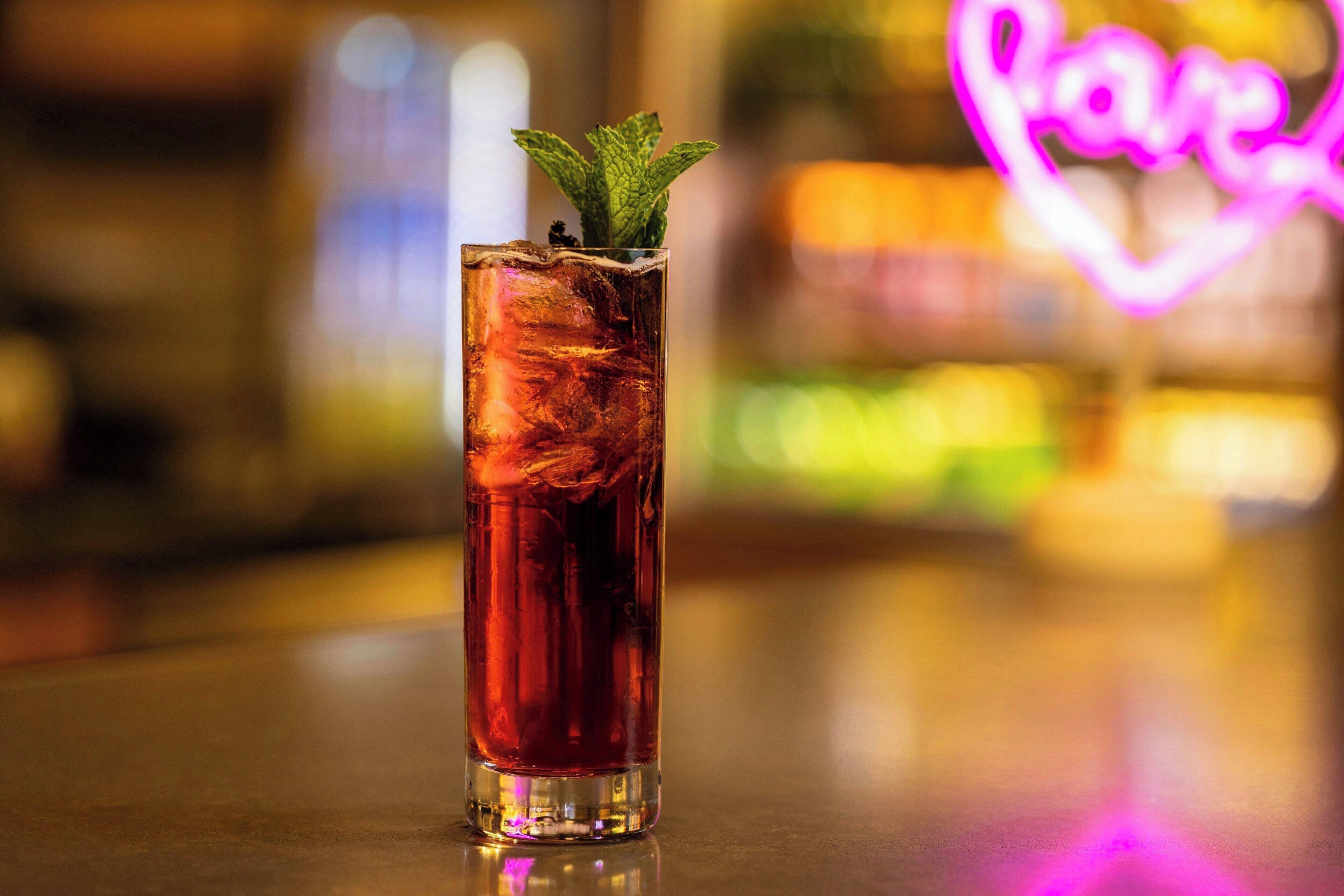A tall glass with a dark liquid and ice cubes, garnished with a mint leaf, on a reflective surface.