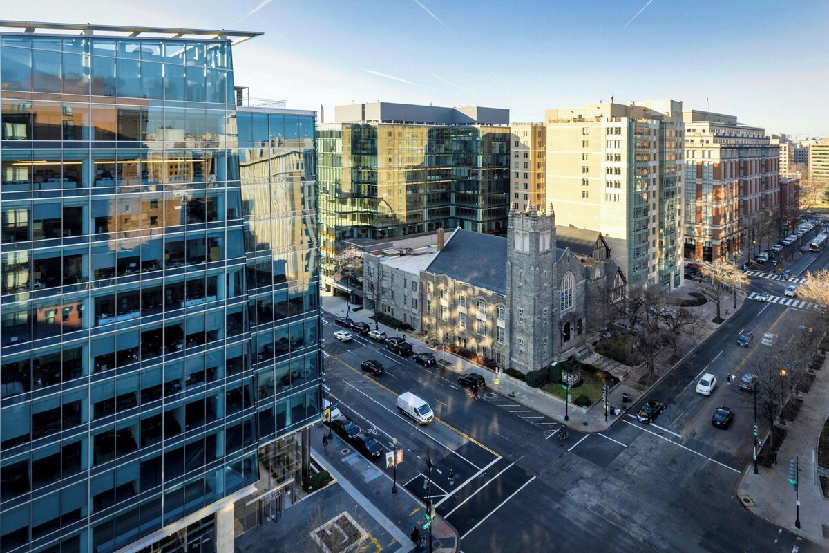 A modern building with a glass facade, a historic building with a stone facade, and a street with cars and a pedestrian crossing.