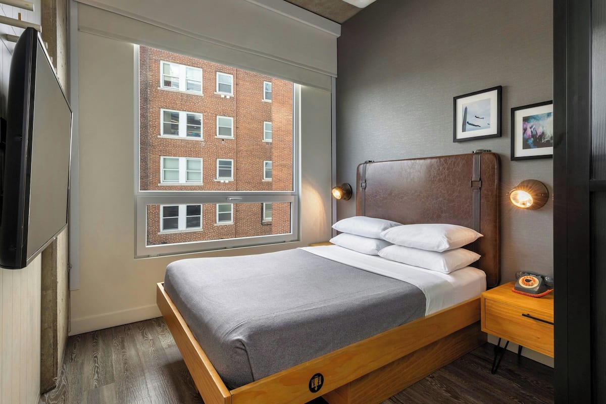 A modern hotel room with a large window, a bed with a wooden frame, a bedside table with a lamp, and a wall-mounted TV.