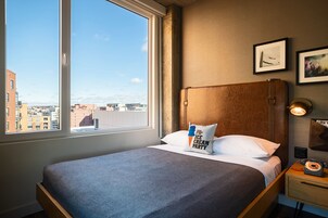 A hotel room with a large window, a bed with a blue bedspread, a pillow with a text design, a nightstand with a rotary phone, and framed pictures on the wall.
