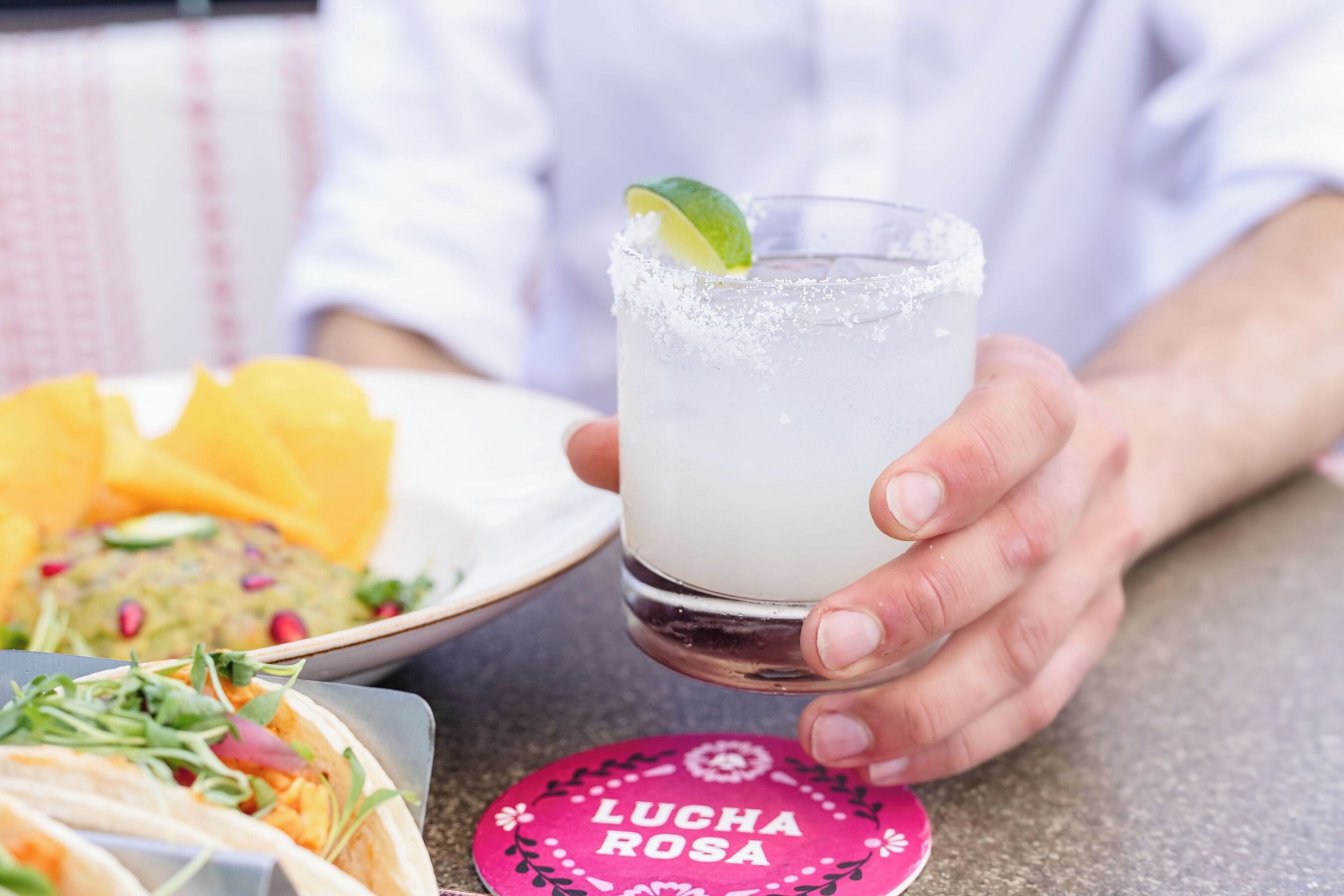 Une personne tenant un verre à margarita avec une tranche de citron vert, une assiette de tacos et un dessous de verre sur lequel est inscrit le texte « LUCHA ROSA ».