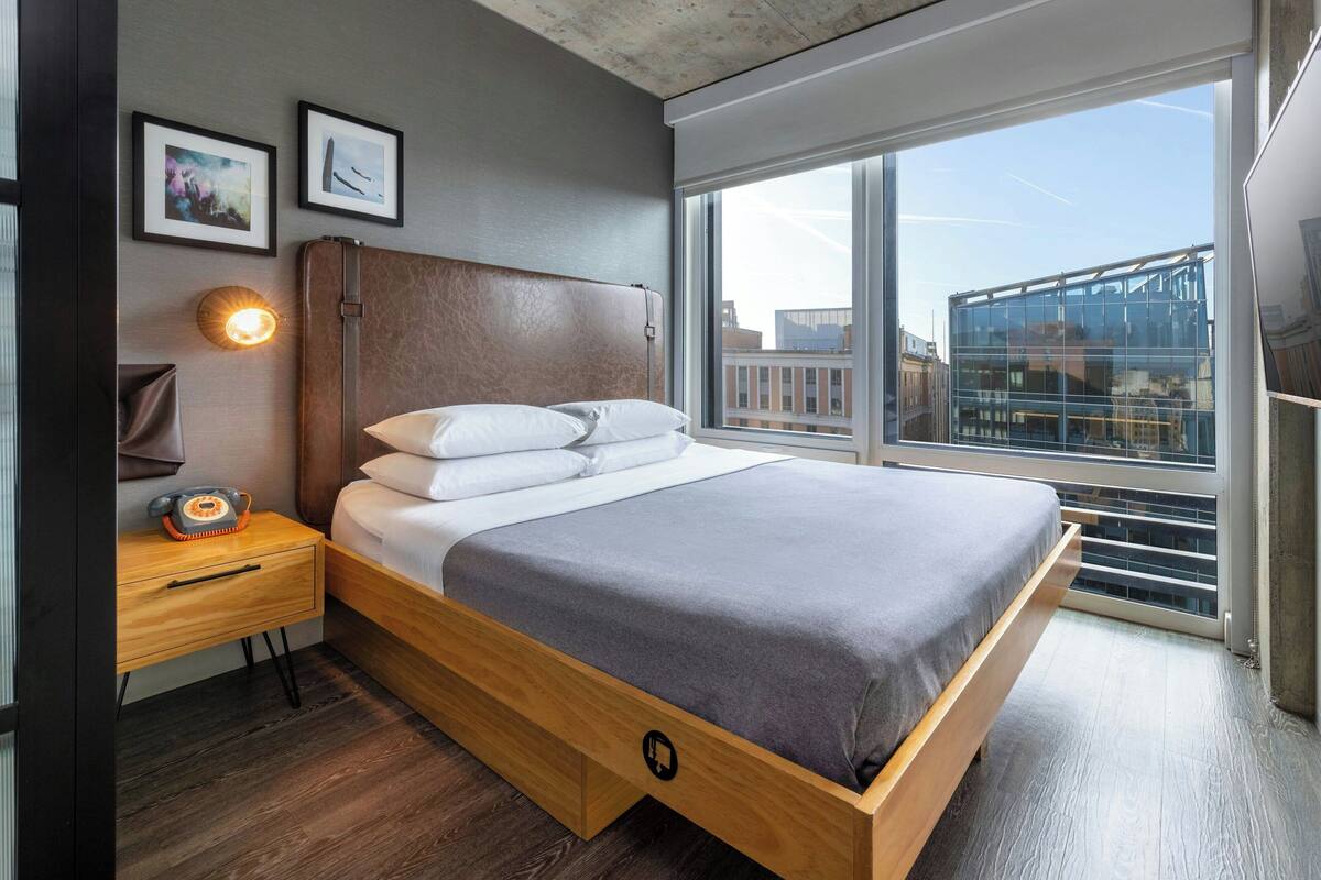 A modern bedroom with a wooden bed, a nightstand with a lamp, and a large window offering a city view.