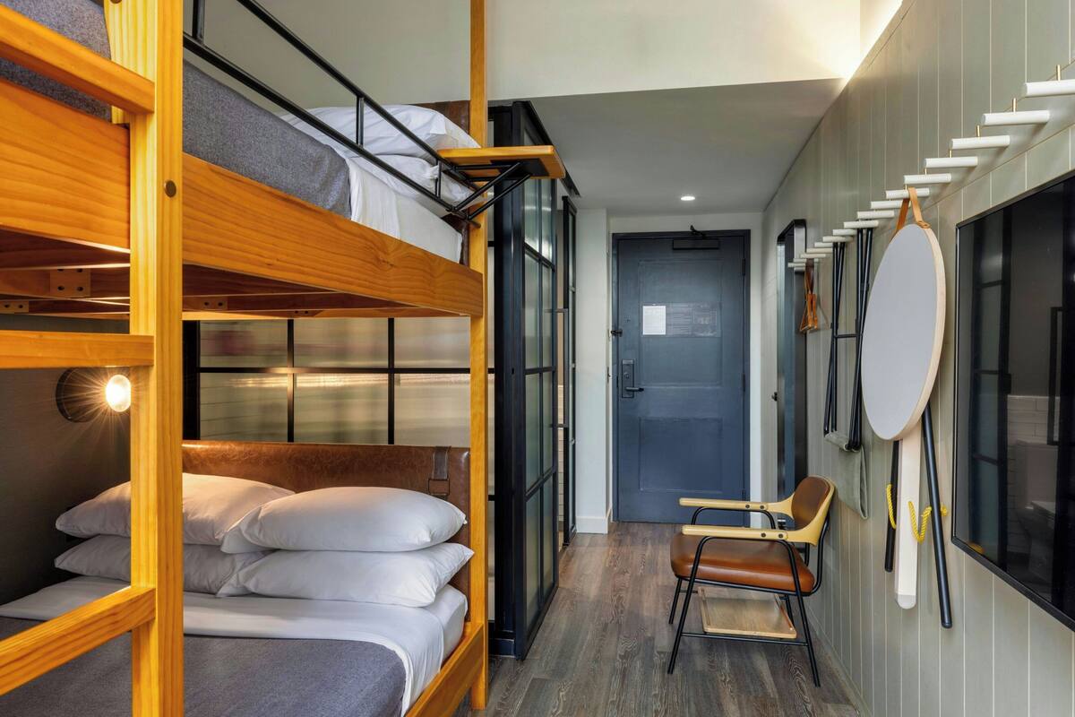 A compact hotel room with a bunk bed, a small seating area, and a large window.