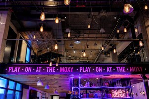 Baarialue, jossa on neonvalomainos, jossa lukee: ”PLAY ON AT THE MOXY”, sekä toinen neonvalomainos, jossa lukee: ”BABE”.