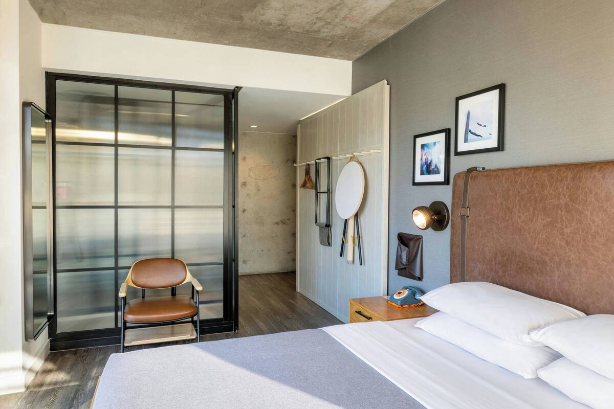 A modern hotel room with a large bed, a chair, a wardrobe, and a mirror.