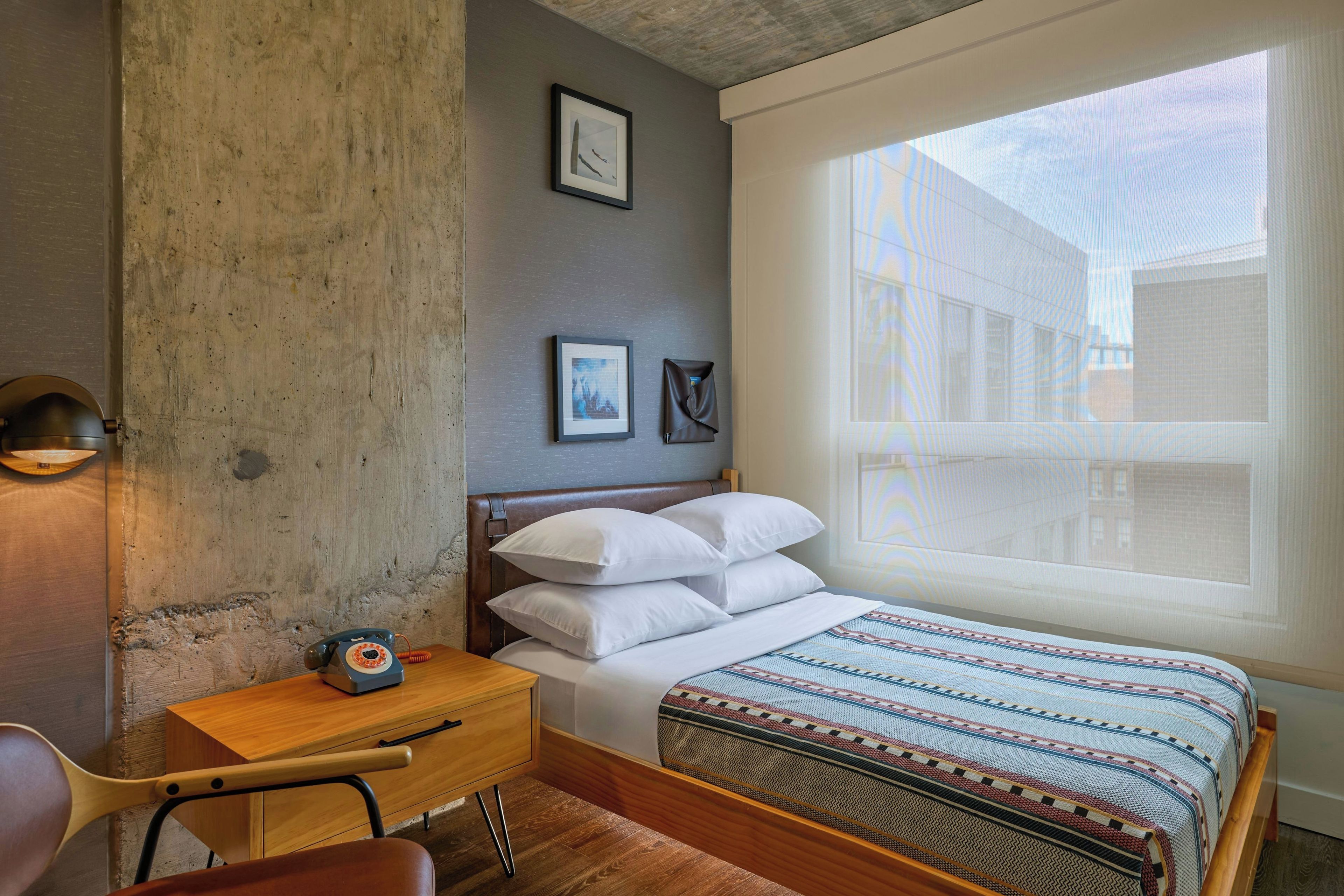A bedroom with a bed, a desk, a chair, a telephone, and a window with a view of buildings.