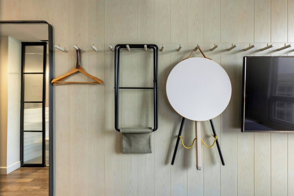 A room with a wooden wall, a coat rack, a hanging coat, a small table with a stool, and a flat-screen TV.