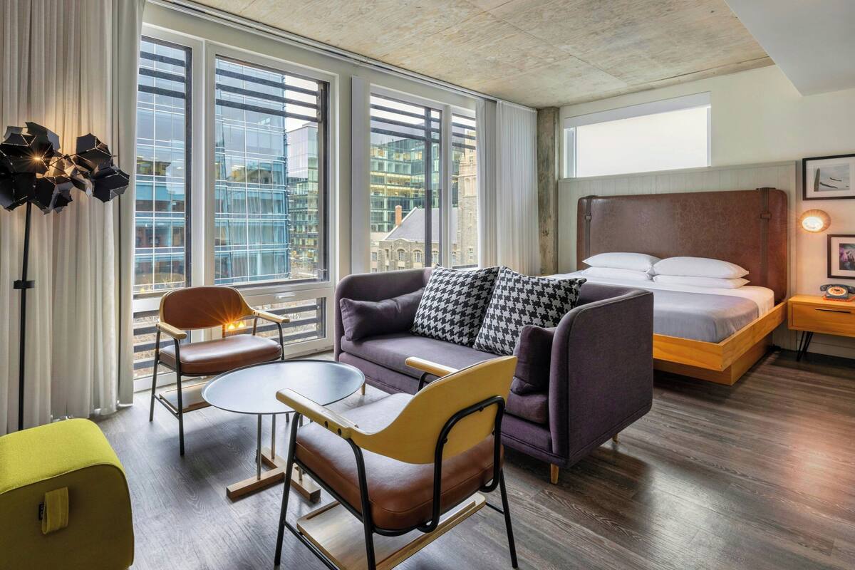 A modern hotel room with a sofa, a small table, a bed, and large windows offering a city view.