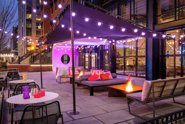An outdoor seating area with tables and chairs, a fire feature, and a covered seating area with cushions. - Moxy Washington DC Downtown (Washington)