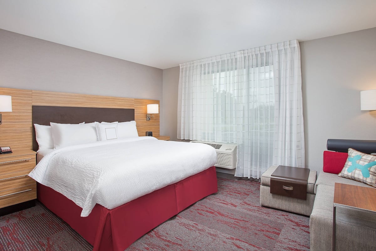 A hotel room with a large bed, a bedside table, a nightstand, a small sofa, a red carpet, and a window with sheer curtains.