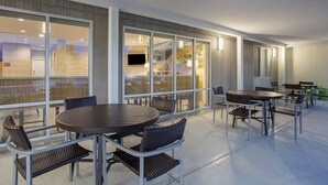 Un área de comedor al aire libre con mesas redondas y sillas de mimbre, ventanas amplias con vistas y un techo plano.