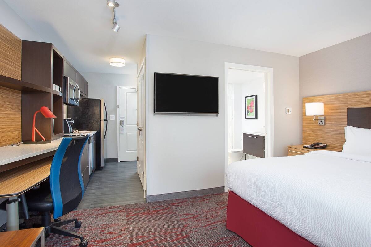 A hotel room with a bed, desk, chair, TV, and a small bedside table.