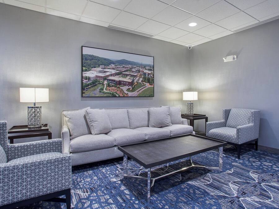 A modern hotel room with a gray sofa, patterned armchairs, a coffee table, and a large landscape painting on the wall.