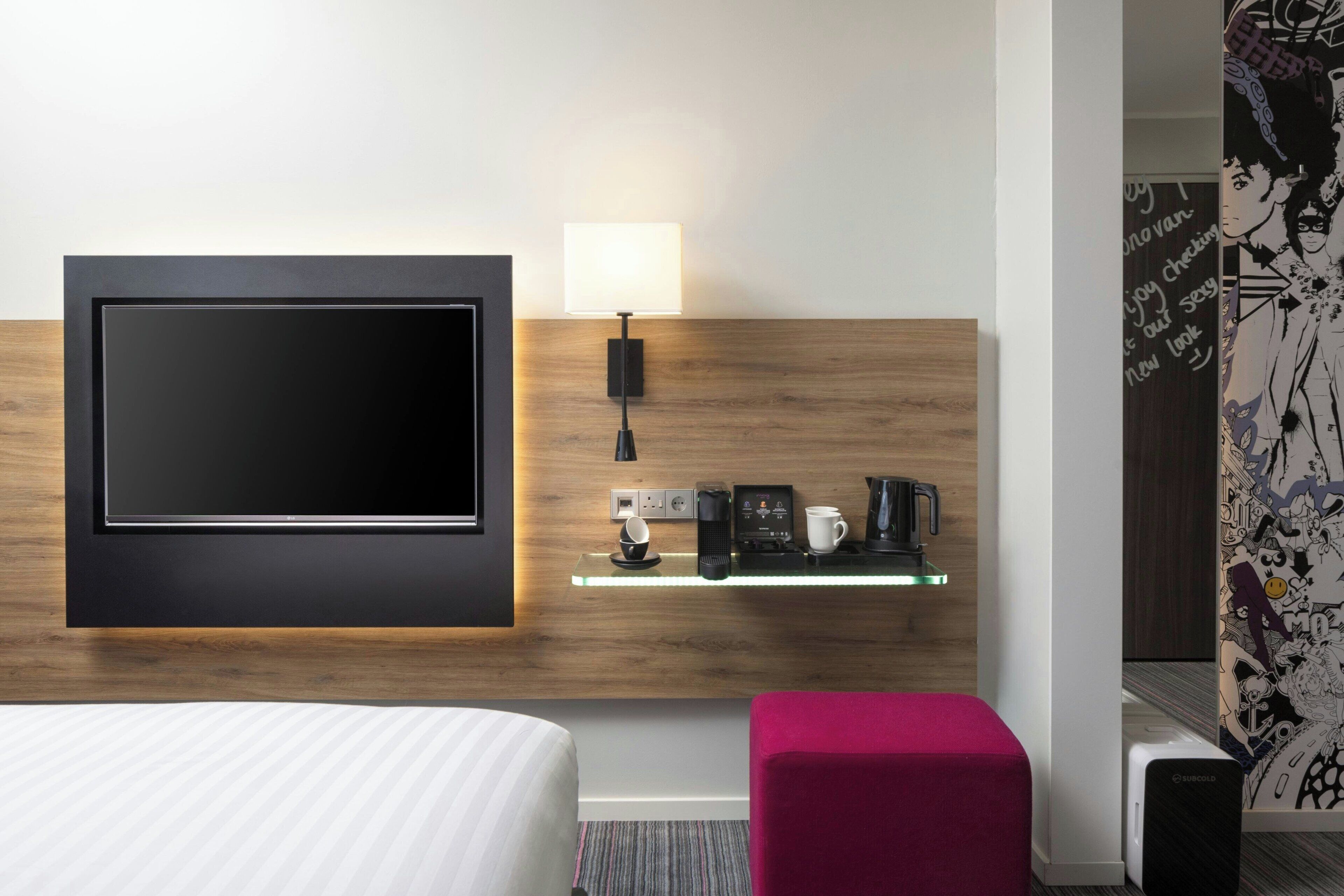 A modern hotel room with a large flat-screen TV mounted on a wooden wall, a bedside table with a lamp, a coffee maker, and a small shelf with a cup and a phone.