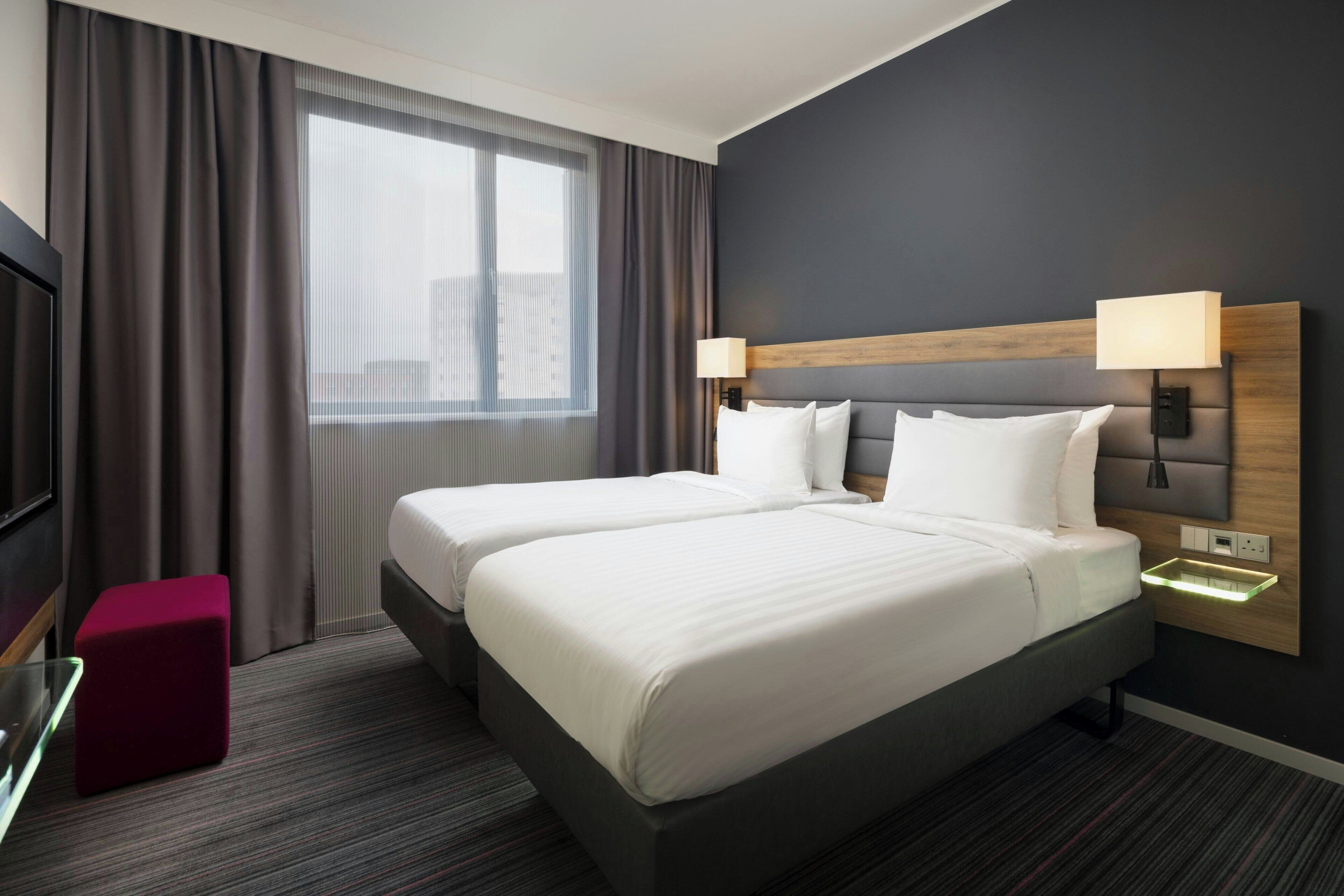 A hotel room with two beds, a TV, a red ottoman, and a window with curtains.
