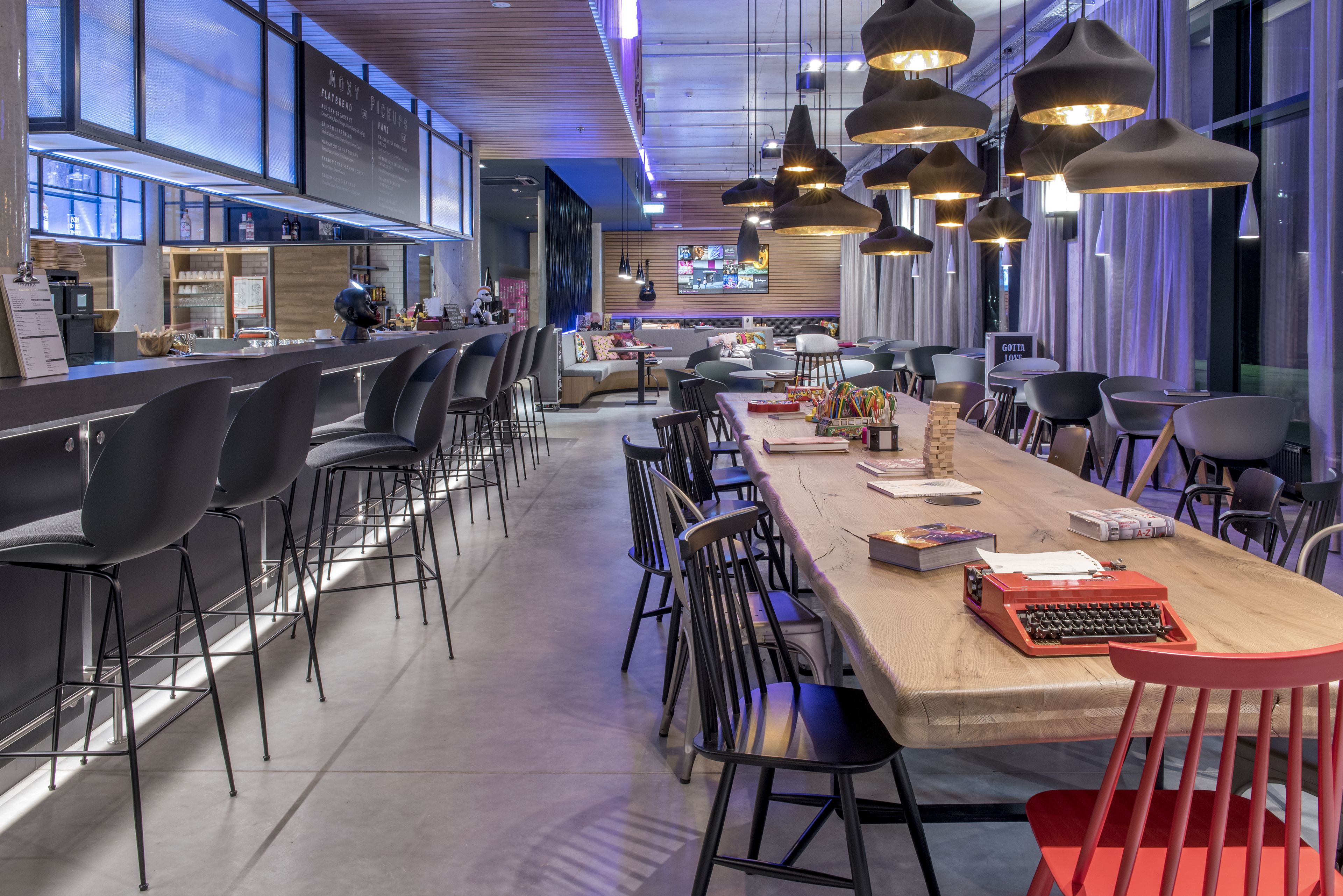 Un restaurant moderne doté d’une longue table conviviale, de chaises noires et rouges, et d’un éclairage suspendu.