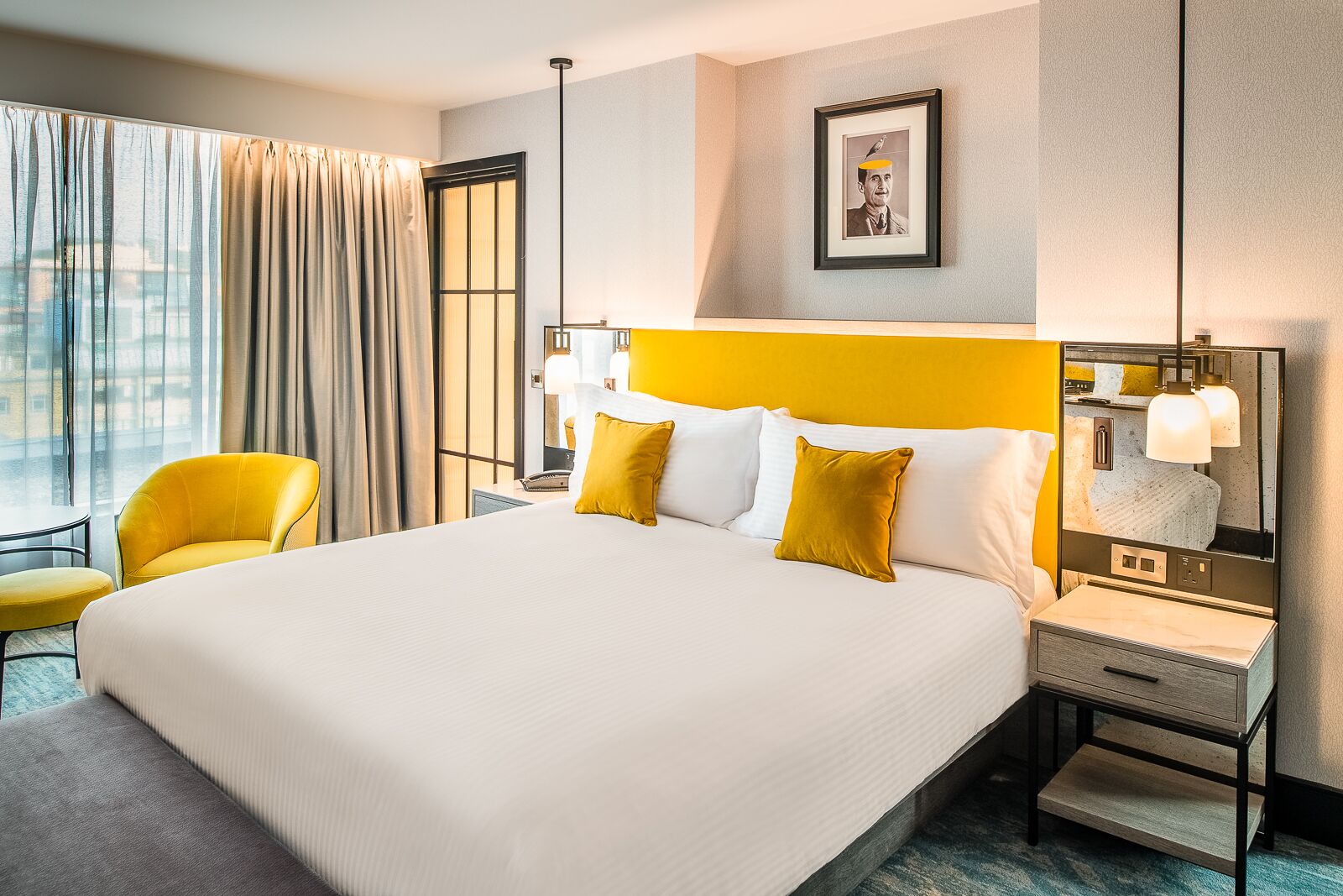 Une chambre d’hôtel avec un grand lit, deux fauteuils jaunes, une table de chevet avec une lampe et un tableau encadré au mur.