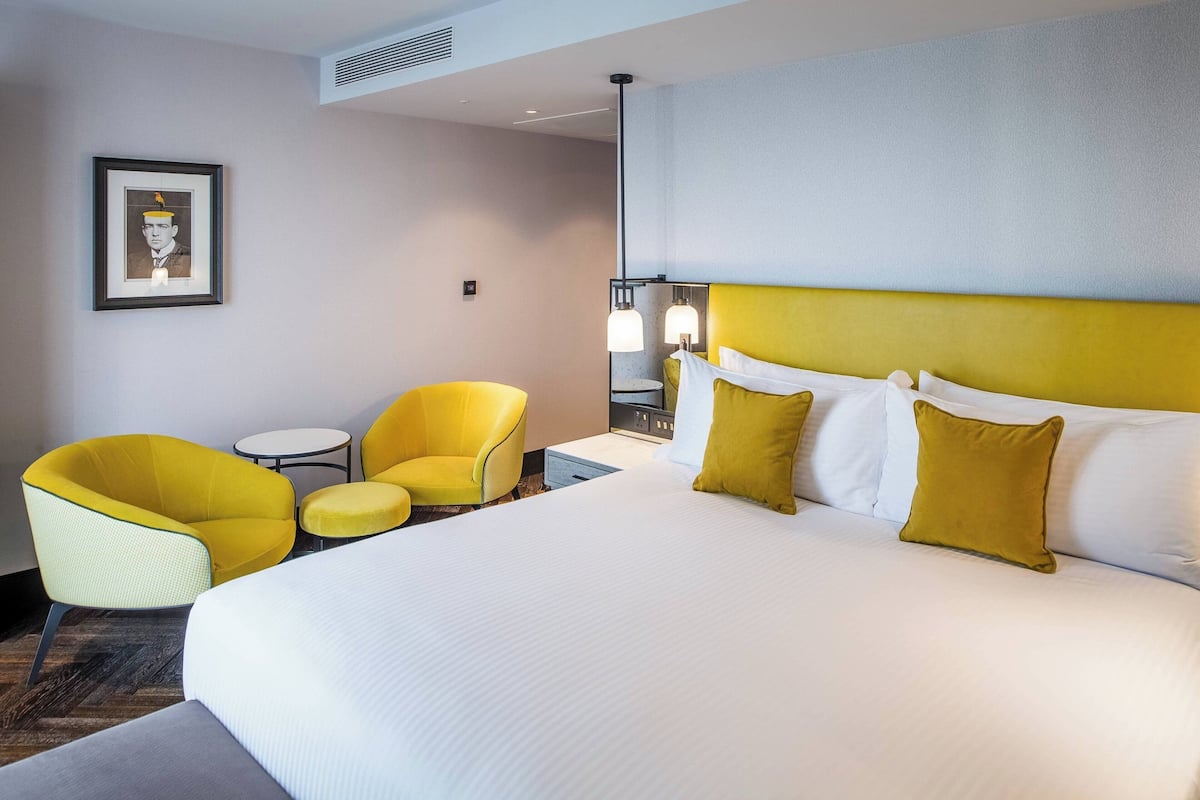 A hotel room with a large bed, two yellow armchairs, a small table, and a framed picture on the wall.