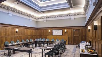 Una sala conferenze con un lungo tavolo, sedie, uno schermo per proiezioni e un angolo buffet con frutta e bevande.