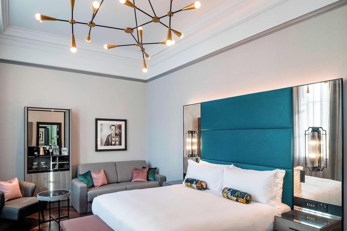 A modern hotel room with a large bed, a seating area with a sofa and chairs, a minibar, and a large mirror.