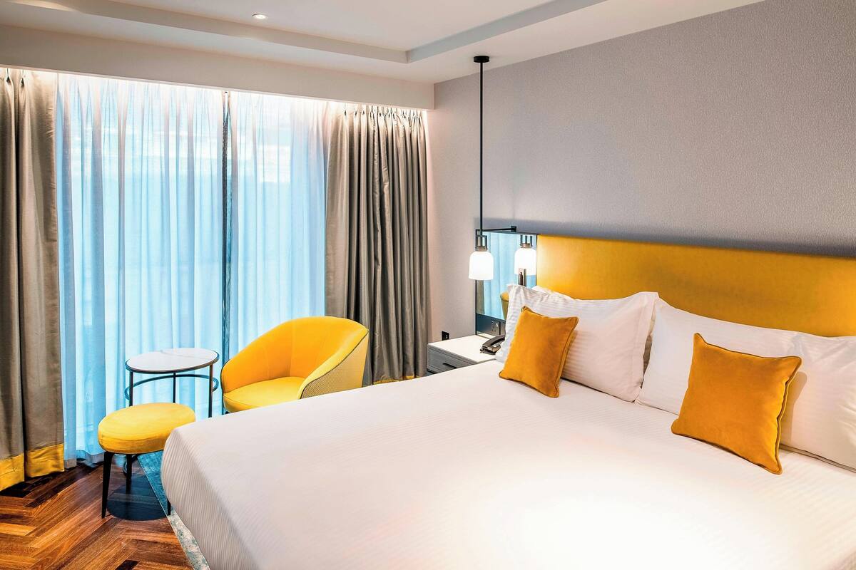 A hotel room with a large bed, a small table, a chair, and a nightstand with a lamp.
